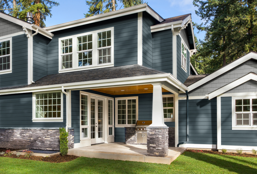 This blue and grey home has white trim and stone accents. A concrete patio area has a grill on it, and green grass is planted in the backyard.